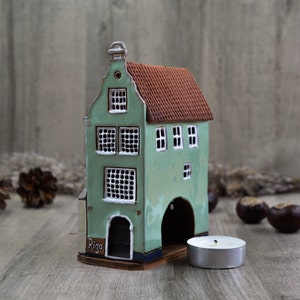 Könnte beinhalten: Ein grünes Keramikhaus mit rotem Dach und einem weißen Torbogen. Das Haus hat ein Schild mit der Aufschrift "Riga" und ist ein Teelichthalter. Ein silbernes Teelicht steht vor dem Haus.