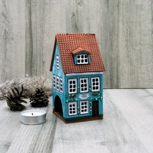 Può includere: Una statuetta di casa in ceramica blu e marrone con finestre bianche e un tetto marrone. La casa ha un cartello che dice "1221" ed è decorata con viti verdi e cervi marroni.