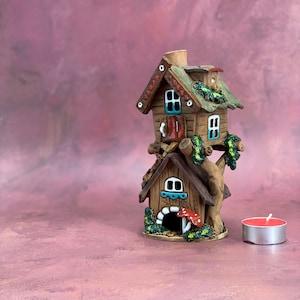 May include: A ceramic fairy house with a brown roof, green trim, and a red door. The house is perched on a tree trunk with a smaller house underneath. A small red candle is in the foreground.