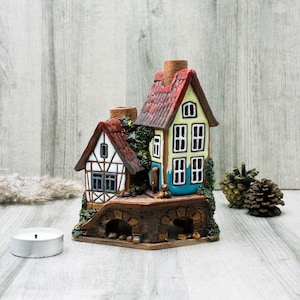 May include: A ceramic house candle holder featuring two houses, one white with a red roof and the other yellow with a red roof, joined by a brown bridge. The houses have windows and doors and are adorned with green ivy.