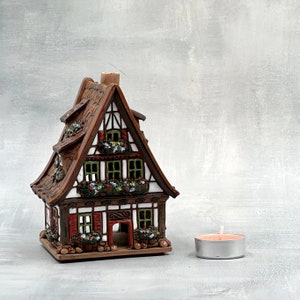 Peut inclure: Un bougeoir en céramique en forme de maison avec un extérieur brun et blanc, des accents rouges et une cheminée. Le bougeoir est décoré de fleurs vertes et blanches et contient une petite bougie.