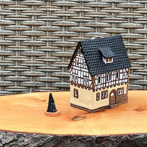 Peut inclure: Une maison miniature en céramique avec un toit noir, des bordures blanches et marron, et une façade beige. Un cône d'encens noir est posé sur un petit porte-encens rond marron. La maison est sur une surface en bois.