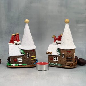 May include: Two decorative miniature houses with white conical roofs and brown brick-like walls. The houses have red chimney stacks with Santa hats and the word "RIGA" on the side. A small red candle sits in front of the houses.