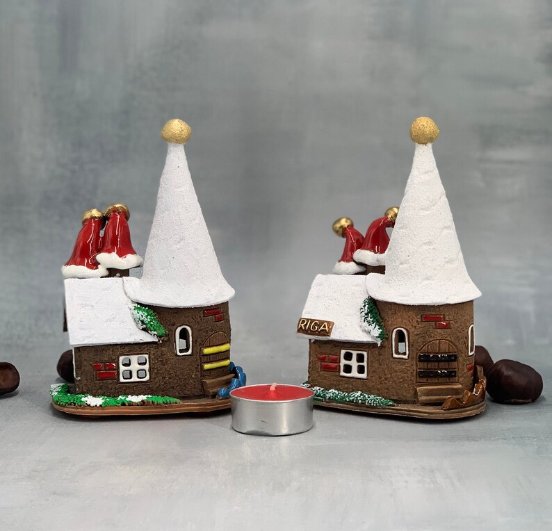 May include: Two decorative miniature houses with white conical roofs and brown brick-like walls. The houses have red chimney stacks with Santa hats and the word "RIGA" on the side. A small red candle sits in front of the houses.