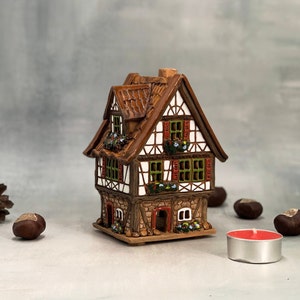 Può includere: Un bruciatore di incenso in ceramica a forma di casa con tetto marrone, pareti bianche e persiane rosse e verdi. La casa ha una base in pietra ed è decorata con fiori.