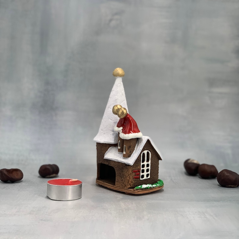May include: A brown ceramic Christmas church with a white steeple and snow-covered roof. A small figure in a red coat is on the roof. A red tea light candle sits in front of the church. Chestnuts are scattered around the church.