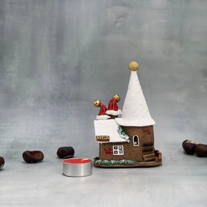 May include: A ceramic Christmas house with a white steeple and a brown base. Two Santa hats sit on the roof. The word "RIGA" is printed on the front. A red tea light candle and chestnuts are in the foreground.