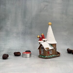 May include: A decorative Christmas house figurine with a white snow-covered roof and red Santa hats. A small red candle sits in front of the house. The house is brown with white trim and a gold ball on top.