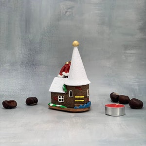 May include: A decorative miniature house with a white conical roof and a gold finial. The house has a brown brick facade, a red roof, and a small green bush. A lit red candle and several chestnuts are in the foreground.