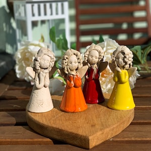 Op de afbeelding: Vier keramische engelbeeldjes met vleugels, elk met een andere gekleurde jurk: wit, oranje, rood en geel. De engelen staan op een hartvormige houten basis.