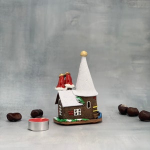May include: A miniature ceramic house with a white conical roof and two red chimney stacks. The house has a brown facade with a small window and door. A small red candle sits in front of the house, and several chestnuts are scattered around.