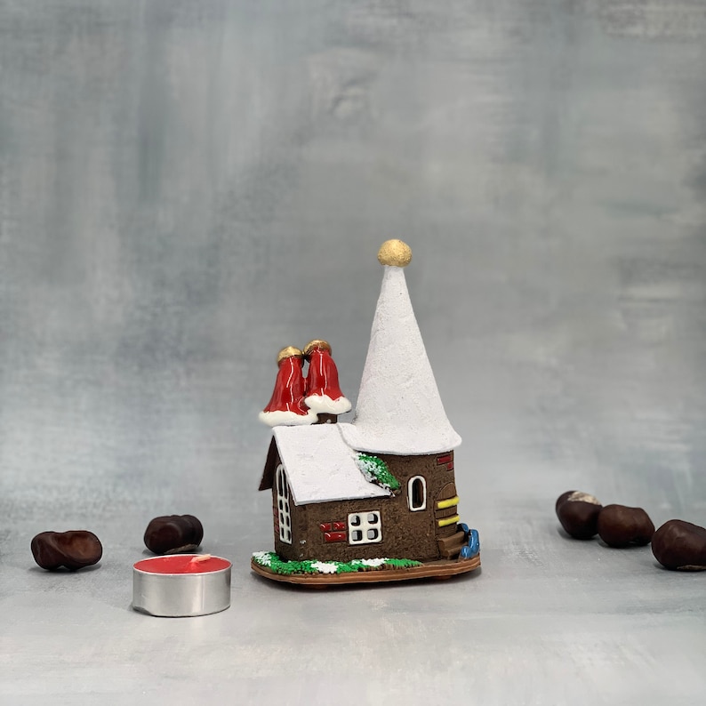 May include: A miniature ceramic house with a white conical roof and two red chimney stacks. The house has a brown facade with a small window and door. A small red candle sits in front of the house, and several chestnuts are scattered around.