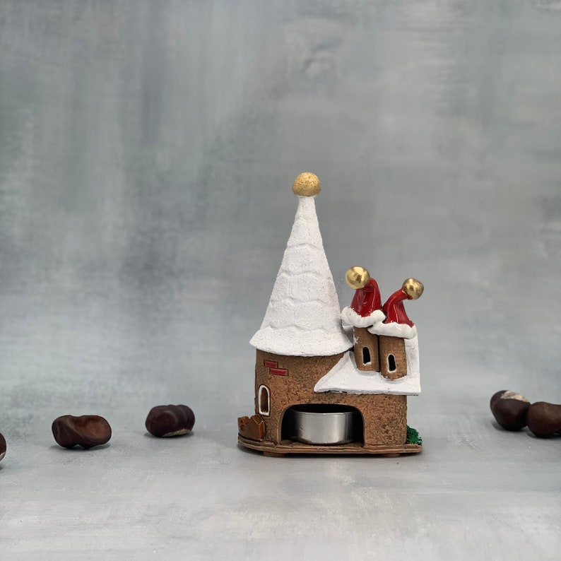 May include: A ceramic Christmas village candle holder. The brown structure has a tall, white, snow-covered steeple and two chimneys with Santa hats. A small tea light sits inside the base. Chestnuts are scattered around the base.
