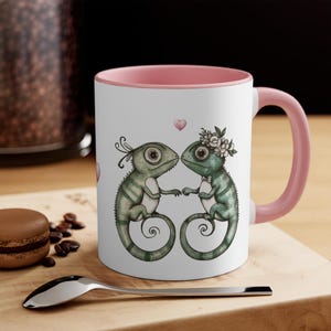 Pode incluir: Uma caneca de cerâmica branca com uma borda e uma alça rosa apresenta uma ilustração caprichosa de dois camaleões verdes com coroas florais, de mãos dadas e olhando um para o outro. Os camaleões estão cercados por corações rosas.