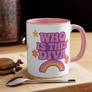 May include: White ceramic mug with a pink interior and handle. The mug features the text "WHO IS THIS DIVA" in a retro font, along with star and flower graphics, and a rainbow design. A spoon and a macaron are in the foreground.