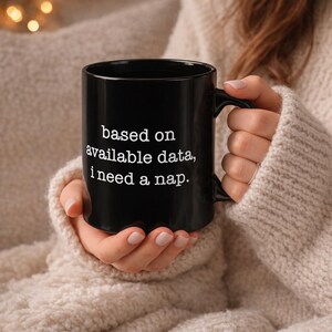 以下が含まれることがあります： 「based on available data, I need a nap」の白い文字が入った黒いセラミックマグ。マグカップはカーブしたハンドルが付いており、ベージュのセーターを着た人が持っています。