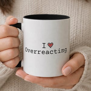 Puede incluir: Taza de cerámica blanca con interior y asa negros. La taza presenta el texto "I ❤️ Overreacting" en negro, con un símbolo de corazón rojo. La taza es sostenida por una persona que lleva un suéter beige.