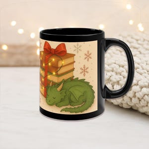 May include: Black ceramic mug featuring a whimsical illustration of a sleeping green dragon next to a stack of books tied with a red bow and fairy lights. The background has snowflakes and a cream color.