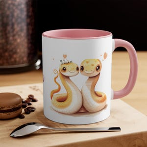 Può includere: Una tazza in ceramica bianca con un bordo e un manico rosa. La tazza presenta un'illustrazione di due serpenti di cartoni animati con cuori e fiori. I serpenti sono gialli e hanno occhi neri.