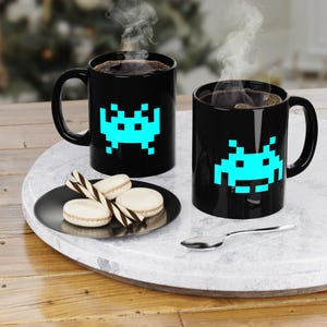 May include: Two black coffee mugs with a pixelated blue alien design. The mugs are on a white marble tray with a black plate of macarons and a silver spoon. Steam rises from the coffee.