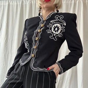 May include: Black jacket with white embroidered detailing and gold buttons. The jacket has a unique design with a high collar and long sleeves. The outfit includes black and white striped pants.