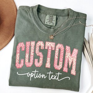 Może przedstawiać: Oliwkowo-zielona koszulka z napisem "CUSTOM" w różowych literach z patchworku. Poniżej słowa "CUSTOM" znajduje się fraza "option text" w kursywie. Złoty naszyjnik i brązowy kapelusz również znajdują się na zdjęciu.