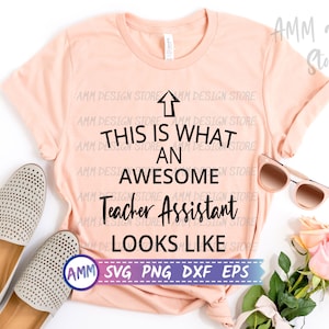May include: A pink t-shirt with the text "This is what an awesome Teacher Assistant looks like" in black lettering. The text is centred on the shirt. The shirt is lying flat on a white surface with a pair of beige shoes and a pair of sunglasses.
