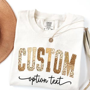 以下が含まれることがあります： フローラルプリントの文字で「CUSTOM」と、筆記体で「option text」と書かれたクリーム色のTシャツ。ゴールドのネックレスと茶色の帽子も写っています。