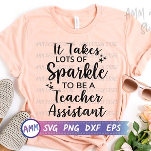May include: A peach-coloured t-shirt with the text "It Takes Lots of Sparkle to Be a Teacher Assistant" in black. The text is surrounded by small stars. The shirt is laid flat on a white surface with a pair of shoes, sunglasses, and pink roses.