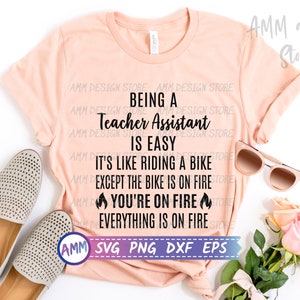 May include: A pink t-shirt with the text "Being a Teacher Assistant is Easy. It's like riding a bike except the bike is on fire. You're on fire. Everything is on fire." The text is in black and there are flame emojis on either side of the text "You're on fire".