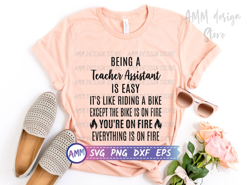 May include: A pink t-shirt with the text "Being a Teacher Assistant is Easy. It's like riding a bike except the bike is on fire. You're on fire. Everything is on fire." The text is in black and there are flame emojis on either side of the text "You're on fire".