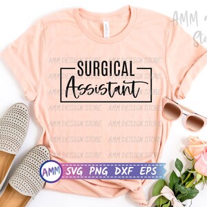以下が含まれることがあります： ピーチ色のTシャツに、黒い長方形の中に「SURGICAL Assistant」という文字が書かれています。Tシャツは白い背景に置かれ、ベージュの靴、サングラス、ピンクのバラが置かれています。