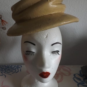 Peut inclure: Un chapeau de couleur beige de style vintage avec un bord incurvé est posé sur une tête de mannequin blanche. Le mannequin a les yeux fermés avec de longs cils noirs, des lèvres rouges et des joues rosées. Le chapeau semble être fait d'un matériau rigide.