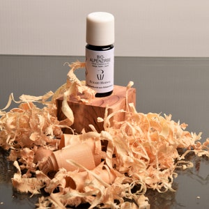 Puede incluir: Una pequeña botella blanca con una etiqueta negra que dice "BIO ALPENZIRBE" y "ECKART HIPP" en un bloque de madera rodeado de virutas de madera.