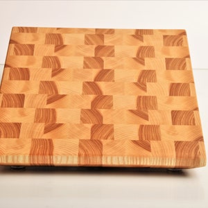 Può includere: Un grande tagliere quadrato in legno con un motivo a scacchiera. Il tagliere è in legno chiaro e ha una finitura naturale. È appoggiato su una superficie bianca.