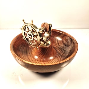 May include: A wooden bowl with a polished finish, featuring a brass ship's wheel nutcracker. The bowl has a rich, warm wood grain pattern. The nutcracker is positioned in the center of the bowl.