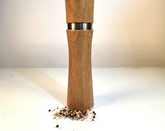 Könnte beinhalten: Eine hohe, zylindrische Pfeffermühle aus Holz mit einer glatten, hellbraunen Maserung. Die Mühle verfügt über ein markantes horizontales silbernes Metallband, das den oberen Mahlabschnitt vom Hauptkörper trennt, mit verstreuten Pfefferkörnern.