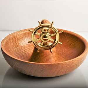 May include: A wooden bowl with a gold-colored ship's wheel centerpiece. The bowl is made of light brown wood and has a smooth, curved design. The ship's wheel is centered and adds a nautical theme to the decorative bowl.