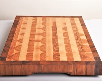 May include: A large, rectangular cutting board made of wood. The board has a light brown center with a dark brown border. The center is made of a patterned wood, while the border is made of a solid wood.