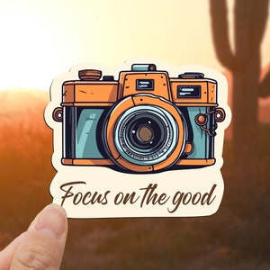 May include: A sticker featuring a vintage camera illustration with the text "Focus on the good".