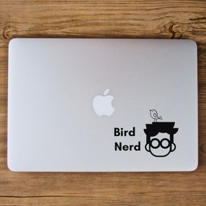 May include: A black and white sticker on a silver laptop. The sticker features a cartoon character with glasses and a bird on their head. The text "Bird Nerd" is below the character.