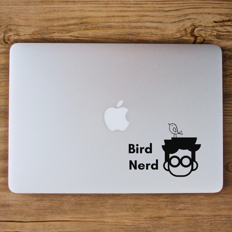 May include: A black and white sticker on a silver laptop. The sticker features a cartoon character with glasses and a bird on their head. The text "Bird Nerd" is below the character.