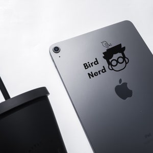 May include: A grey iPad with a black sticker that says "Bird Nerd" and a cartoon image of a person with a bird on their head. The Apple logo is visible on the back of the iPad.