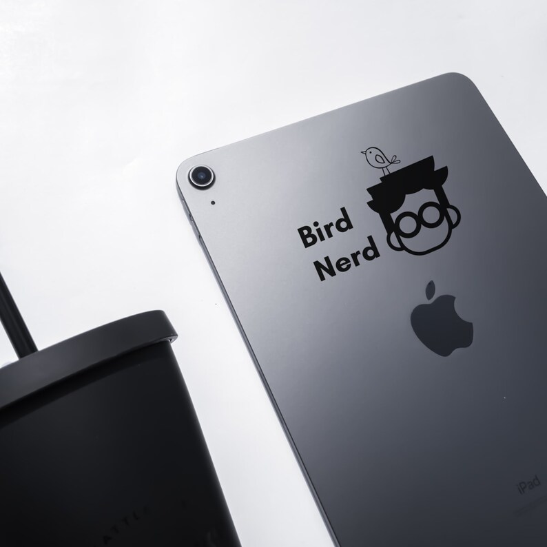 May include: A grey iPad with a black sticker that says "Bird Nerd" and a cartoon image of a person with a bird on their head. The Apple logo is visible on the back of the iPad.