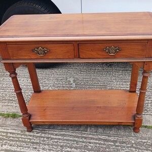 May include: A wooden console table with two drawers and a lower shelf. The table has a light brown finish and brass drawer pulls.