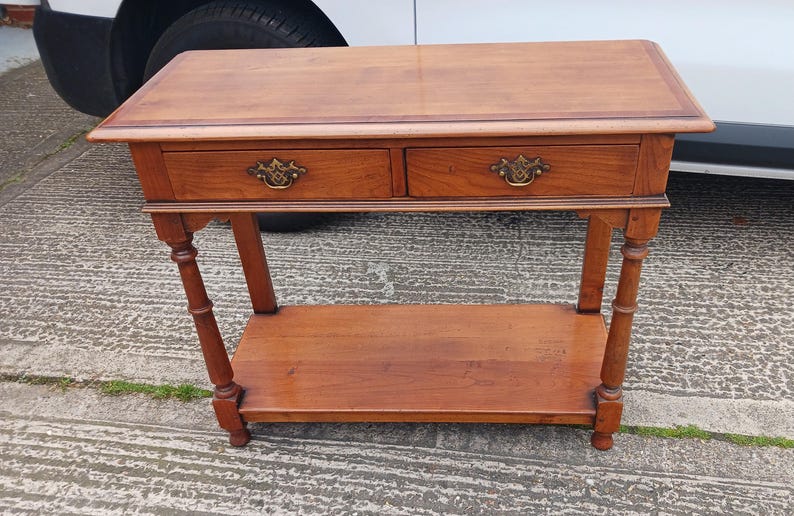 May include: A wooden console table with two drawers and a lower shelf. The table has a light brown finish and brass drawer pulls.