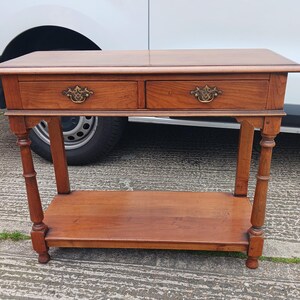 May include: A wooden console table with two drawers and a lower shelf. The table has a light brown finish and brass hardware.