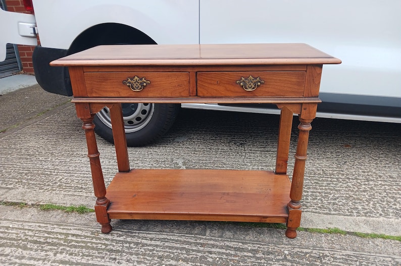 May include: A wooden console table with two drawers and a lower shelf. The table has a light brown finish and brass hardware.