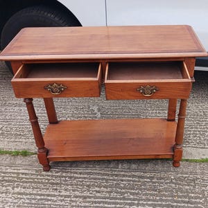 May include: A wooden side table with two drawers and a lower shelf. The table has a light brown finish and brass hardware.