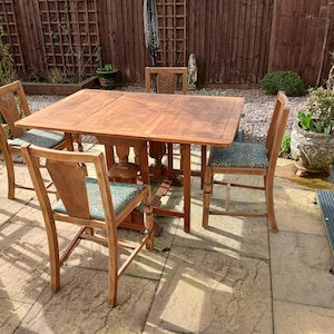 May include: A wooden dining table and four chairs set. The table is rectangular with a light brown finish and a pedestal base. The chairs have wooden frames and patterned seat cushions. The set is placed on a stone patio, suitable for outdoor dining.